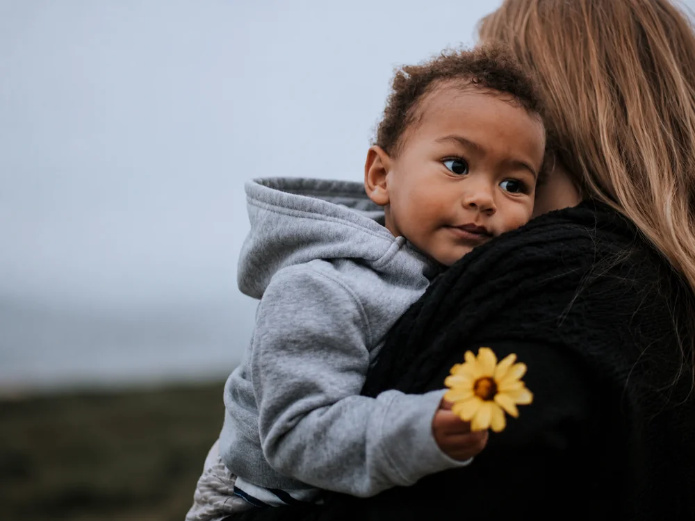 A parent carrying their smiling child, who is holding a yellow flower